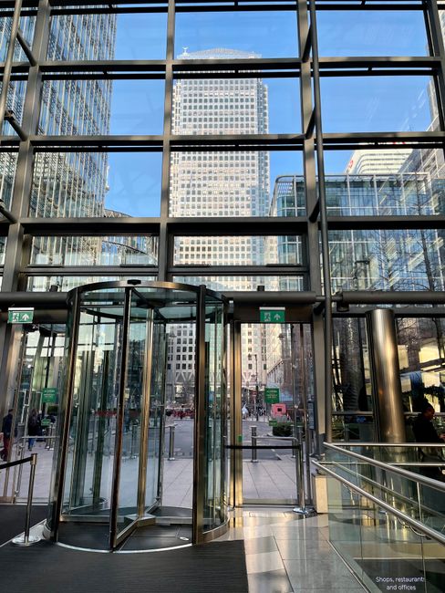Interior view of a modern commercial building with large floor-to-ceiling glass windows revealing an urban cityscape with high-rise buildings under a clear blue sky. The entrance features a curved gla