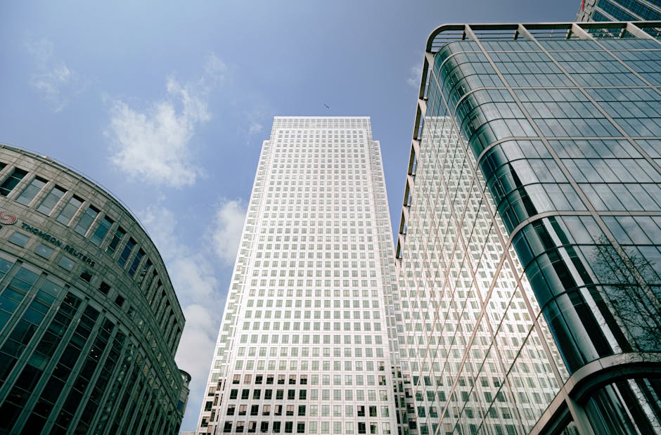 Interior view of a modern commercial building with large floor-to-ceiling glass windows revealing an urban cityscape with high-rise buildings under a clear blue sky. The entrance features a curved gla